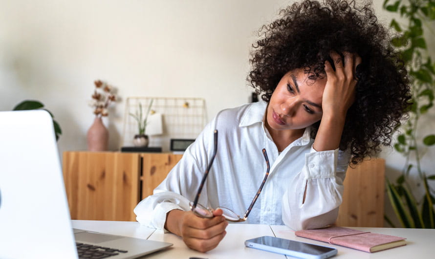 Woman at a desk rests her head on her hand, holding glasses, with a laptop, phone, and notebook on the desk.